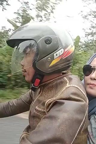 A passenger wearing sunglasses and a light blue hijab is riding on the back of a motorcycle. The passenger is wearing a brown leather jacket. The motorcycle is being driven by someone wearing a dark helmet. The background shows trees and vegetation, indicating an outdoor setting. The video is a close-up shot of the passenger and the driver's helmet.