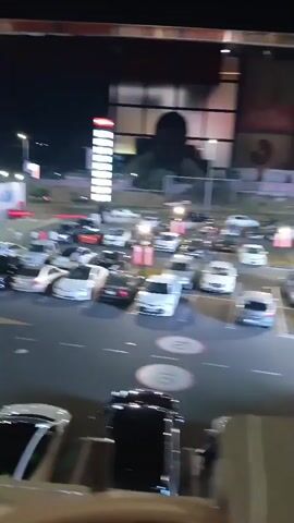 The video shows a parking lot at night. Many cars are parked, some are white, black, and gray. There is a tall red and white sign with lights in the background, and a building with glass windows. There are some street lights in the parking lot. In the foreground, there is a railing. The video is shaky and the lighting is dim. No people are clearly visible.
