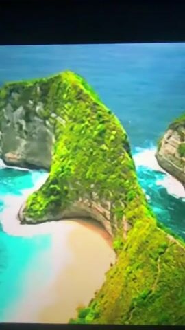 An aerial drone shot captures a dramatic coastal cliff formation covered in lush green vegetation. The cliff juts out into the ocean, resembling the head and neck of a dinosaur. Waves crash against the rocks at the base of the formation, and a small sandy beach is visible in the cove. The water is a vibrant turquoise color. The scene is serene and natural, with no visible human activity.