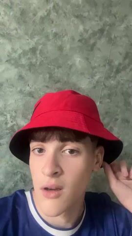A young person wearing a red bucket hat and a blue shirt is looking directly at the camera. The person's right hand is raised and touching the brim of the hat. The background is a textured green wall. The video appears to be a selfie.