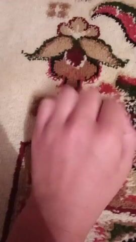 A close-up shot shows a person's hand interacting with a patterned carpet. The hand, with fair skin, is in motion, and its fingers are seen extending, flexing, and making contact with the carpet's surface. The carpet features a floral or decorative motif in muted colors, primarily cream, with accents of red, green, and brown. The lighting suggests an indoor environment, casting soft shadows. The camera appears to be held at a slightly downward angle, focusing on the hand and the carpet.