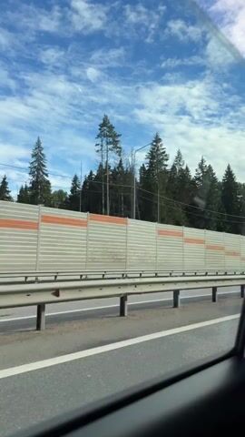 A moving shot from inside a vehicle, looking out the passenger side window. The view shows a highway with a sound barrier wall made of corrugated metal panels, some with orange horizontal stripes. Beyond the wall, a dense forest of coniferous trees is visible. A cell tower stands tall among the trees on the left side of the frame. The sky above is blue with scattered white clouds. The road surface appears wet in places. The vehicle is moving forward at a moderate speed, indicated by the passing of the sound barrier and trees.