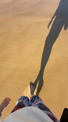 A first-person perspective video shows a person walking on a sandy beach. The person's bare feet are visible, as are their legs wearing shorts with a graphic print. The shadow of the person, elongated by the low sun, stretches across the sand. Waves gently lap at the shore, washing over the person's feet. The water is clear with foam from the waves. The sand is a light brown color. The sun is casting a warm glow.