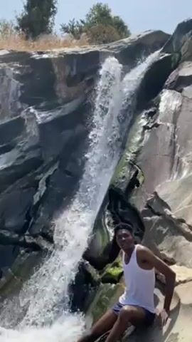 A young man wearing a white tank top and dark shorts is sitting near a waterfall on a bright, sunny day. He is partially on a large rock, with the waterfall cascading down behind him. The rocks are dark and jagged, with some green moss. He points at the camera and smiles broadly. The waterfall rushes down the rocks and splashes around him. There are some trees visible at the top of the waterfall.