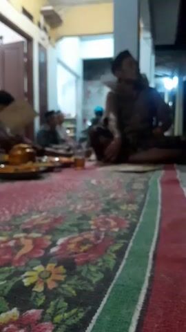 The video shows an indoor gathering of several men, likely a religious or social event, taking place in Panjalin Kidul, Indonesia. The setting appears to be a home, with a traditional rug covering the floor. The room is dimly lit, suggesting it is nighttime or evening.

Several men are seated on the floor, some in discussion or prayer. In the foreground, a low table is laden with food and drinks, including what appears to be a golden-brown mound of food, possibly rice or a traditional dish, and several glasses of tea. The men are dressed in traditional attire, including peci (caps), which are commonly worn by Muslim men in Indonesia.

One man, in the right of the frame, is more prominent. He is seated with his legs crossed, looking upwards, and appears to be engaged in conversation or contemplation. Other men are visible in the background, some in discussion, others seemingly listening or observing. The overall atmosphere seems to be one of community and shared experience. The floral pattern on the rug is vibrant and colorful, contrasting with the subdued lighting of the room. There is no visible text in the media.
