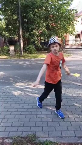 A young boy wearing an orange t-shirt with a soccer ball graphic and a black and white checkered hat is standing on a paved sidewalk. He is also wearing black pants and blue sneakers. The boy is holding a yellow object, possibly a toy, and appears to be spinning it. The background shows a street, trees, and houses, suggesting a suburban or residential area. The sunlight indicates it is daytime. The boy's expression is difficult to discern, but his posture suggests he is engaged in playing with the object. The scene is set in Kamionka, Poland.