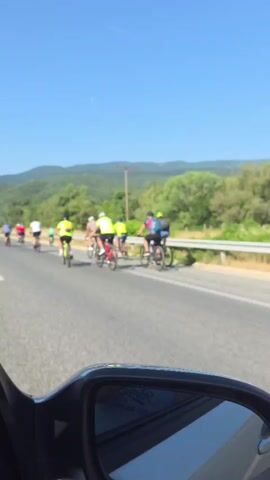 A video shows a group of cyclists on a road, as seen from a vehicle's side mirror. The cyclists are wearing various colored shirts and helmets, riding bicycles of different types. They are cycling on the right side of the road, which has a white solid line marking the shoulder. To their right, there's a guardrail and beyond that, green trees and hills. The sky is clear and blue. The video is taken from inside a moving vehicle, with the side mirror visible in the foreground.