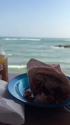 The video shows a lunch consisting of a drink labeled "Papelon" in a glass jar, and a arepa on a blue plastic plate, against the backdrop of the ocean. The arepa is partially wrapped in paper and the filling is visible.  A crumpled white napkin sits beside the plate. The ocean is calm, with gentle waves and visible rocks in the water. The sky is clear and blue.