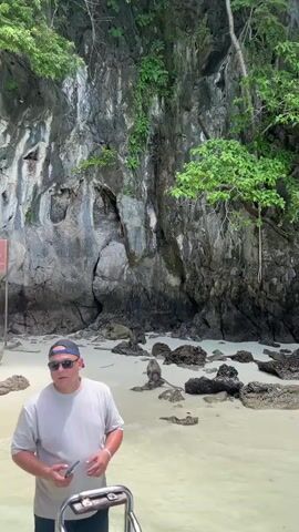 This is a video taken from a boat, showing a beach with a rocky cliff face covered in lush greenery. A man in a light-colored t-shirt, dark shorts, sunglasses, and a baseball cap stands in the foreground, holding a phone. To his left, another man in a patterned shirt and shorts walks along the beach. Further down the beach, a group of people are gathered near a sign. One person is holding a rope. There are also several monkeys visible on the beach. The water is shallow and clear.