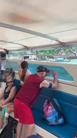 A group of tourists are on a covered boat, looking out at the ocean. The water is turquoise, and there are rocky islands covered in green vegetation in the distance. Several people are taking photos with their phones. One man in a red shirt and red shorts is leaning against the window and holding a phone up to take a picture. A woman in a black and white swimsuit is sitting to his left. A woman with a black baseball cap is standing and taking a photo. Another person in a white hat and white shirt is standing and looking out. A man in a plaid shirt and green hat is also standing and looking out. There is a colorful backpack on the blue bench seat.
