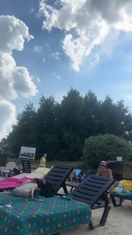 This is a vertical video clip filmed on a beach. The scene is a sandy beach with lounge chairs set up. In the foreground, there are several empty black lounge chairs, one of which is covered with a colorful patterned towel. Behind the lounge chairs, several people are visible. One person in a yellow bikini is standing near the water. Another person is sitting in a lounge chair, facing away from the camera. A third person in a patterned headscarf and swimsuit is sitting in a lounge chair and appears to be looking down. A child's inflatable toy is on a small table. In the background, there is a dense line of tall green trees and a clear blue sky with fluffy white and gray clouds. The sun is partially obscured by clouds, suggesting it is midday or early afternoon. The weather appears to be warm and pleasant for a beach outing. The overall mood is relaxed and leisurely. There is a sign on an easel to the left of the frame, but the text is too small to be legible.