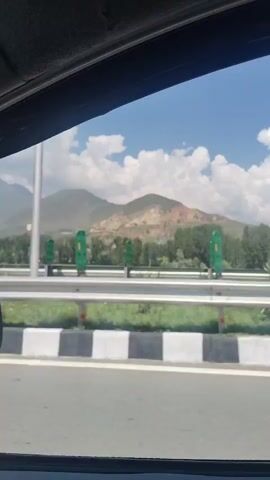 The video shows a view from inside a vehicle moving along a highway. The highway has a divided lane with guardrails and a grassy median. Beyond the median, there are trees and mountains in the background. The sky is partly cloudy. The video captures the scenery passing by as the vehicle travels.