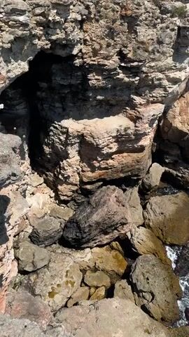 The video pans across a rocky coastline with the turquoise ocean. In the foreground, large, rough-hewn rocks are scattered along the shoreline, with waves crashing against them. As the camera moves upwards and to the right, it reveals sheer, weathered cliffs made of light-colored stone. The water below is a deep, vibrant blue-green, with white foam from the waves breaking on the rocks. In the distance, the ocean stretches out to meet a clear blue sky. At the very end of the video, a few small figures can be seen standing on the clifftop, observing the view. There are no other discernible objects or people in the video.