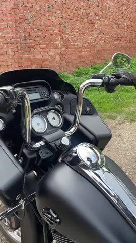 This is a close-up, eye-level video of the handlebars and front section of a matte black motorcycle. A person wearing a black glove is operating the clutch lever. The motorcycle has chrome handlebars, mirrors, and gauges. A digital display is visible above the gauges. The scene is set outdoors with a red brick wall and green grass in the background. The lighting suggests it is daytime, possibly overcast given the lack of harsh shadows. The overall mood is neutral, focusing on the mechanics and operation of the motorcycle. No discernible text is visible in the video, though a logo that appears to be Harley-Davidson is present on the fuel tank. The location is identified as Oktyabr'skii, Russia.