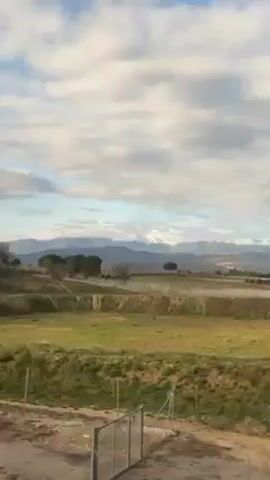 The video shows a landscape seen from a moving vehicle. In the foreground is a rural field area, changing from yellowish-green to brown. In the distance are mountains, partially snow-covered and under a cloudy sky. The camera moves past trees.