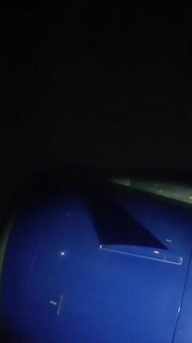 The video shows a close-up, dimly lit view from the window of an airplane at night. The blue engine casing of the plane dominates the foreground, with a dark shadow cast across it. In the distance, faint lights suggest a city or landing strip. The scene appears to be during a flight, possibly just before landing or during takeoff.