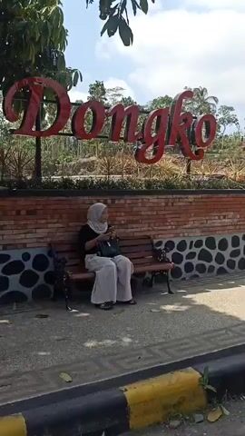 The video shows a street scene with a brick wall and a large sign that reads "ongko" in red letters. A woman in a light-colored headscarf and clothing is sitting on a bench to the left of the sign. Behind the wall, there are trees and some buildings. Further down the street, several people are riding motorcycles. One motorcycle has a license plate that reads "AG 3249 YCE". There is also a red and white circular sign on a stand in the road. The sky is blue with some clouds.
