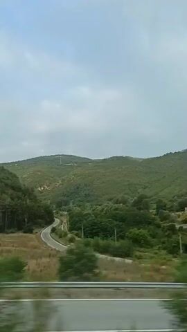 The video shows a scenic view from a moving vehicle on a cloudy day. The landscape is characterized by rolling green hills covered in dense vegetation and trees. A winding asphalt road cuts through the valley, disappearing into the distance. The foreground features a paved area with a small, white building that appears to be a rest stop or service facility. The vehicle is moving along a road with a metal barrier separating it from the surrounding environment. The vegetation on the hills is a mix of trees and shrubs, with some drier, grassy areas visible closer to the road. The sky is overcast with clouds, suggesting a diffused light or potentially an impending change in weather. There are no people visible in the frame.