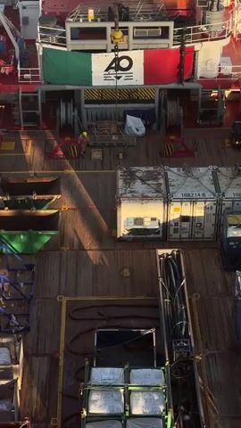 An aerial view shows the deck of a ship. A person in an orange safety suit is walking on the wooden deck. Several shipping containers are stacked on the deck, some with visible serial numbers. The Italian flag is draped across the upper deck of the ship, alongside a banner with the number "40" and other symbols. A crane hook is visible above the banner. Various equipment, including pipes and storage containers, are also present on the deck. The sun casts long shadows across the deck, indicating a low sun angle.