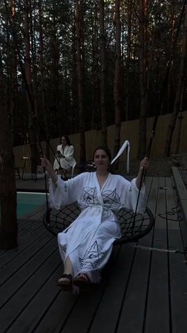 A young woman wearing a white robe with geometric designs sits on a circular swing suspended by ropes. She is in the center of the frame, holding onto the ropes with both hands, and her legs are extended forward. Her robe has wide sleeves and is cinched at the waist with a belt. She is wearing sandals. In the background, slightly out of focus, another woman in a white robe is seated on a chair. The scene is set outdoors in a wooded area with tall, dark trees. A wooden deck surrounds the swing and the seating area. The lighting suggests it is dusk or early evening, with shadows cast across the deck. The overall atmosphere appears relaxed and serene.