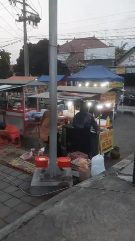 The video shows an outdoor market scene. There are several food stalls set up under canopies. People are visible sitting and standing around the stalls, some appear to be customers and others vendors. The ground is paved with interlocking bricks, and there are some plants and trees in the background. The lighting suggests it is either early morning or late evening. One stall has a sign that reads "SEDAP AYAM MIE TELOR". A pole with electrical wires is prominent in the foreground.