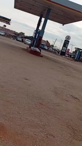 The camera pans across a dusty, unpaved area of land, showing a gas station canopy in the foreground. Other elements in the scene include vehicles, power lines, small commercial buildings with red roofs, and a water tank. The sky is overcast with dark, heavy clouds, though a patch of sunlight manages to break through. There is movement of pedestrians and motorcycles visible.