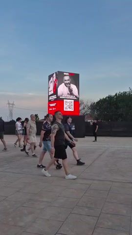 A group of people walk across a paved area in front of a large LED screen displaying images of two men and a QR code. The sky is clear and blue with some clouds. The screen is mounted on a cube-shaped structure. Trees and fencing are visible in the background. The people are casually dressed and appear to be attending an outdoor event or festival.