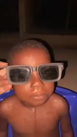 A close-up shot shows a young, dark-skinned child sitting in a blue plastic tub. The child is wearing oversized, black sunglasses. Water droplets are visible on the child’s face and upper body. A hand is seen briefly placing the sunglasses on the child. The child looks up and smiles, revealing a few small teeth. The background is dark and nondescript. The audio includes someone speaking in a language other than English, followed by the child repeating some of the words.