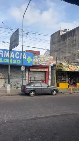 The camera faces a street in Argentina and slowly zooms in on a business, on whose storefront sign Lionel Messi is shown smiling and holding hot dogs, next to the words "SUPER PANCHOS SANDWICHES DE MILANESA EMPANADAS BEBIDAS." To the left of the business is a pharmacy called Alonso. A grey car is parked in front of the business. Two people walk across the frame.