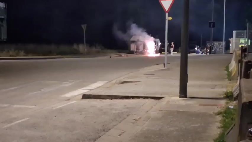 The video captures a nighttime scene on a street in Mas Aliu, Spain. The primary activity is the detonation of fireworks, which are creating bright flashes of light and plumes of smoke. Several people are present, observing the display.

In the background, a small white structure, possibly a kiosk or a portable building, is illuminated by the fireworks. A white van is parked nearby, and a few individuals are standing around it. Further to the right, a group of people is seated, also watching the event. The overall setting appears to be an urban or semi-urban area, with streetlights and road markings visible.

The time of day is clearly night, indicated by the darkness and artificial lighting. The weather is not explicitly discernible, but the presence of smoke suggests a calm atmosphere. The emotions of the people are not clearly conveyed due to the distance and darkness, but their presence suggests engagement with the fireworks. No visible text is present in the media file.