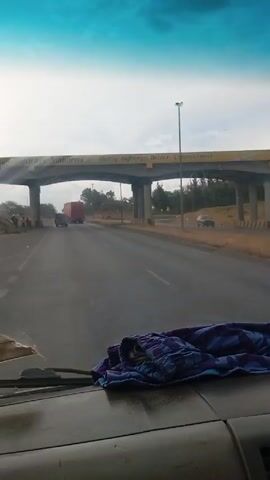 The video, taken from inside a vehicle, shows a highway overpass with the words "National Highways Authority" and "Quality Highways, Better Connections" displayed in yellow lettering. The road is wet, suggesting recent rain. Two trucks and a car are visible on the road in the distance. There are a few people standing on the side of the road to the left. In the foreground, on the vehicle's dashboard, lies a crumpled blue and purple patterned garment. The sky above is mostly cloudy with a patch of blue.