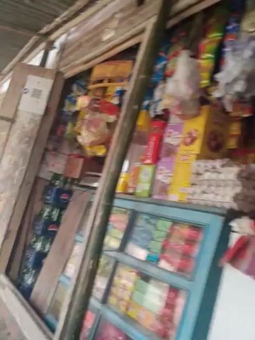A small shop is packed with various items. On shelves, there are stacks of boxes containing snacks like cookies or biscuits in colorful packaging. Bags of chips or snacks are also visible, some hanging and others stacked. In a glass display case, rows of bottled drinks, likely sodas, are neatly arranged. A QR code, possibly for payments, is prominently displayed on the shop's structure. The shop appears to be made of simple materials, with wooden planks and possibly a corrugated metal wall. The overall impression is of a well-stocked, local convenience store.