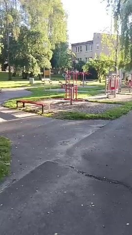 The video shows an outdoor exercise equipment area in a park. The equipment is red and metallic, with some wooden elements. There are multiple pieces of equipment, including what appears to be a leg press machine and a pull-up bar. A red bench is also visible. The ground is a mixture of asphalt and gravel. Tall trees surround the area, and a building can be seen in the background. The lighting suggests it is daytime, possibly morning or afternoon. There are no people visible in the video.
