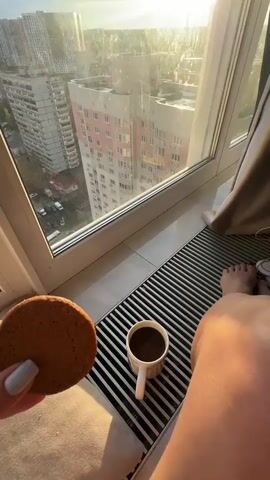 A person is seen indoors, holding a round cookie in their left hand, with a white mug containing coffee placed on a radiator below a large window. Their bare feet and lower legs are also visible next to the radiator. The window offers a view of a cityscape dominated by several tall residential buildings, including one with pinkish-white facades. The sun is low in the sky, either rising or setting, casting bright golden light and long rays across the scene and through the window, which appears to have some smudges or dirt. The sky outside is partly cloudy. The overall atmosphere suggests a quiet moment of enjoying coffee and a snack while observing the city from above, likely in Gavrilkovo, Russia.