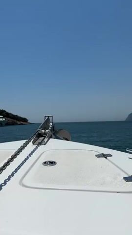 This media file captures a forward-facing view from the bow of a white motorboat sailing on a clear, calm, turquoise sea under a bright, cloudless blue sky. The sun is high, indicating it is midday. On the deck of the boat, a prominent silver anchor is mounted on a roller, with a thick metal chain trailing from it. Two white deck hatches with stainless steel latches are visible.

In the distance, to the left, a lush, green, rocky coastline with scattered trees juts out into the water. To the right, the horizon stretches out, meeting the sea and sky in a seamless line, with a faint landmass visible in the far distance. The water appears tranquil, with gentle ripples reflecting the sunlight. The overall mood is one of peaceful travel and serene enjoyment of a sunny day at sea. There is no visible text in the media file.