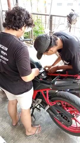 Two individuals are shown working on the rear section of a motorcycle. The individual on the left, wearing a black t-shirt with white text and light-colored shorts, stands with their back to the camera, observing. The individual on the right, wearing a black t-shirt and a black baseball cap, is leaning over the motorcycle, appearing to be manipulating electrical components or wiring under a black plastic cover. The motorcycle has a black fuel tank and red frame and rims. The ground is paved with concrete. In the background, a metal fence and some foliage are visible, along with a blurred image of a vehicle.