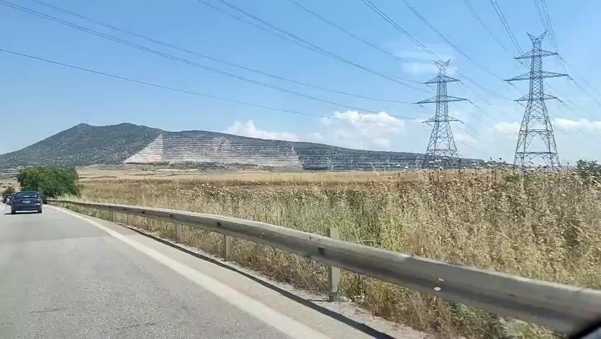 The video shows a driving perspective from a vehicle on a two-lane asphalt road under a clear blue sky. A metal guardrail runs along the right side of the road. To the right of the road is a dry, grassy field. In the distance, a large mountain dominates the left side of the frame, featuring a prominent, multi-tiered white quarry or mine excavation on its slope. Further to the right, multiple tall metal electricity pylons with overhead power lines stretch across the landscape. Several cars are visible on the road, with some moving away from the camera and others in the opposing lane.