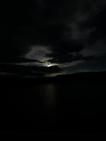 The camera looks out over a body of water, possibly a lake or reservoir. The sky is mostly cloudy, but there is a break in the clouds where the sun shines through, creating a bright reflection on the water. The surrounding landscape consists of hills and some vegetation. The scene is somewhat dark, with a contrast between the bright sunlight and the dark clouds and land. Over the course of the video, the cloud cover shifts slightly, changing the amount of light reflected on the water.