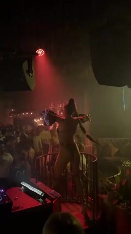 A DJ set is in progress in a dimly lit club. Red and yellow spotlights sweep across the room, illuminating a crowd of people dancing. On a raised platform, a performer in a revealing outfit is dancing and interacting with the audience. The DJ booth is visible in the foreground with turntables and other equipment. The atmosphere appears energetic and festive.