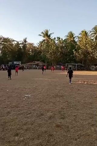 The video shows an outdoor soccer game being played on a dry, grassy field. The sky is clear and bright, indicating daytime. Palm trees and other vegetation form a backdrop to the field. Several players in red and black uniforms are visible on the field, engaged in the game. A soccer goal is set up on one end of the field. Spectators are gathered around the perimeter of the field, watching the match. The overall atmosphere appears to be casual and community-oriented, likely a local game or tournament.