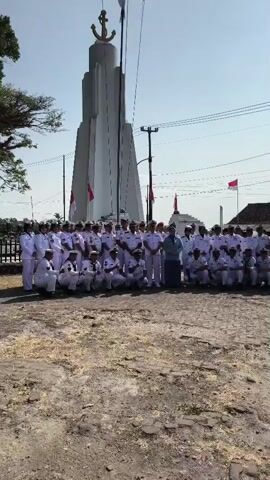 A large group of people, mostly men in white uniforms with dark accents, are posing for a photograph in front of a tall, white monument topped with an anchor symbol. Many are kneeling in the front, while others stand behind them. One person in the center of the group, wearing a blue shirt and dark pants, appears to be directing or interacting with the uniformed individuals. The monument is in a public outdoor space, with a dirt and gravel ground cover. Two Indonesian flags are visible, one on a pole near the monument and another in the background. The sky is clear and bright.