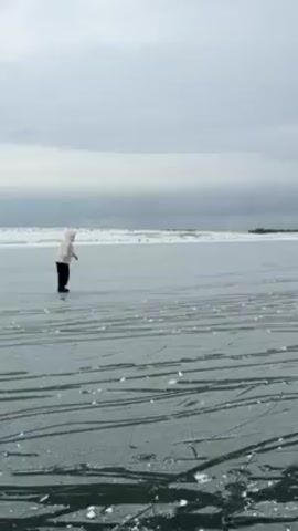 The video shows a person ice skating on a frozen lake or body of water. The person is wearing a light-colored jacket and dark pants, and is seen from the back. The ice surface is marked with numerous skate tracks. In the distance, a thin line of land is visible, possibly with some vegetation. The sky is overcast with a cloudy, gray sky. Later in the video, other skaters can be seen in the distance.