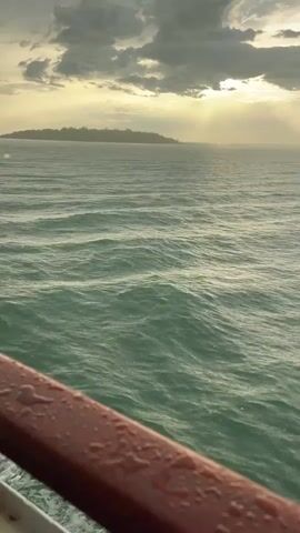 The video shows a view of the ocean from a moving boat. The sun is low in the sky, casting a golden glow through the clouds and onto the water. There is a small island in the distance, covered in trees. The water is a deep green and is choppy with small waves. The railing of the boat is in the foreground, wet with what appears to be rain droplets. The scene suggests a time of day around sunset or sunrise, with dramatic lighting from the sun breaking through an overcast sky. The overall mood is serene and perhaps a little melancholic, with the vastness of the ocean and the distant island creating a sense of isolation. The location is identified as Don Sak, Thailand, a coastal town known for its ferry connections. No text is visible in the video.