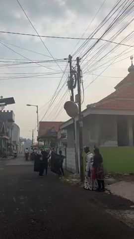 A street scene in what appears to be a village or town. A dirt or asphalt road runs through the middle, lined with houses and buildings on either side. A few people are visible, some walking, others on motorcycles. Several people are gathered in the street, some holding flags or banners. There are also food vendors with carts set up along the roadside. Overhead, a tangle of electrical wires crisscrosses the sky, suspended from utility poles. The sky is overcast, with the sun peeking through the clouds. The overall impression is of a community gathering or a local event.