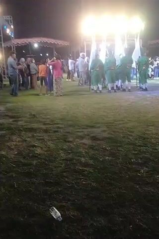 The video captures an outdoor nighttime event with artificial lighting. In the foreground, a patch of grass is visible, with a discarded plastic water bottle lying on it. In the midground, a group of individuals dressed in green uniforms with white hats and holding white flags are standing and marching. They appear to be part of a procession or performance. Behind them, a crowd of spectators is gathered, some standing under tents. The lighting is bright, illuminating the central area of activity. The overall atmosphere suggests a festive or ceremonial gathering.