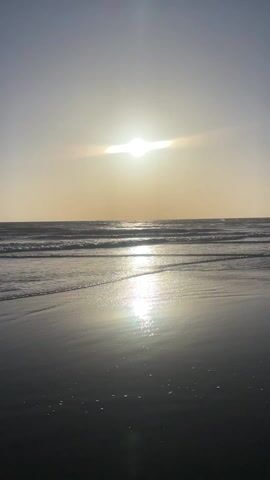 The video depicts a beach scene at sunset. The sun is positioned high in the sky, with a bright glare obscuring its form. The sunlight reflects intensely off the water, creating a shimmering effect on the wet sand. Gentle waves are rolling onto the shore. The overall tone is calm and serene.
