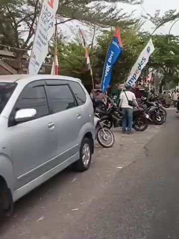 The video shows a street scene with numerous motorcycles parked along the side of the road. Several people are visible, some standing near the motorcycles and others riding them. Flags are displayed, one with blue and red lettering that appears to say "BANK DKI" and another with blue and white lettering that says "SIMPEDES." The environment appears to be an outdoor market or roadside area with trees and buildings in the background. The sky is overcast. The overall impression is of a busy street with a lot of motorcycle activity.