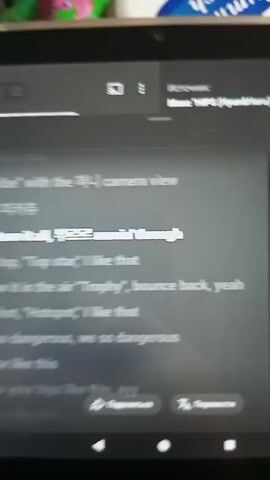 The video shows a close-up of a tablet screen displaying lyrics. The lyrics are in a mix of English and Korean. The screen is lit, and the lyrics are white and highlighted in blue. The song title is "HI-P" and the artist is "Hyun&Haru". The lyrics mention "XX vibe with the camera view", "Haru 피카츄", "turn it all, movin' through", "Top, top, 'Top star', I like that", "Throw it in the air 'Trophy', bounce back, yeah", "Hot, hot, 'Hotspot', I like that", "We so dangerous, we so dangerous", "Dance like this", "Move your hips like this, ayy". There are also icons at the bottom of the screen for sharing and translation. The tablet appears to be resting on a surface, possibly a bed or a table, with blurred objects visible in the background, including a pink and green stuffed toy. The video is recorded from a slightly angled perspective, looking down at the screen.