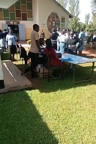 The video shows an outdoor scene with several people and objects. A group of students, dressed in school uniforms, are gathered around a table where a woman is seated, likely handling paperwork or a registration process. A man stands next to her, seemingly engaged in a conversation. Other students can be seen walking and standing nearby. 

The setting appears to be a school or educational institution, indicated by the presence of students in uniforms and a building in the background with a sign or design on it. The area is grassy, with some trees visible. The overall atmosphere seems to be one of activity and organization, possibly related to school registration or some similar event.