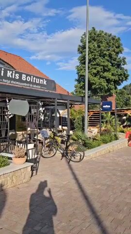 The video shows a street scene in what appears to be a European town. The camera pans from left to right, starting with a close-up of a cafe called "Mi kis Boltunk". The cafe has outdoor seating and a bicycle parked in front. The camera then pans to show more of the street, which is paved with bricks and lined with trees and buildings. There are other cafes and shops visible, as well as people walking along the street. The sky is blue with some clouds, and the sun is shining. The overall impression is one of a pleasant, relaxed atmosphere.